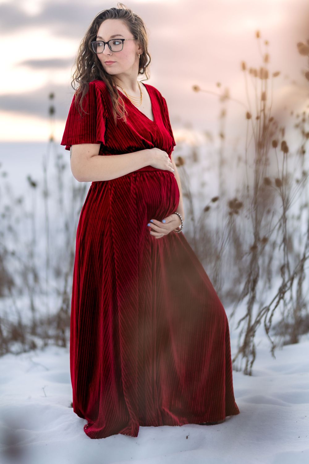 Pregnant woman in elegant red velvet gown holding her baby bump outdoors in a snowy winter landscape at sunset.