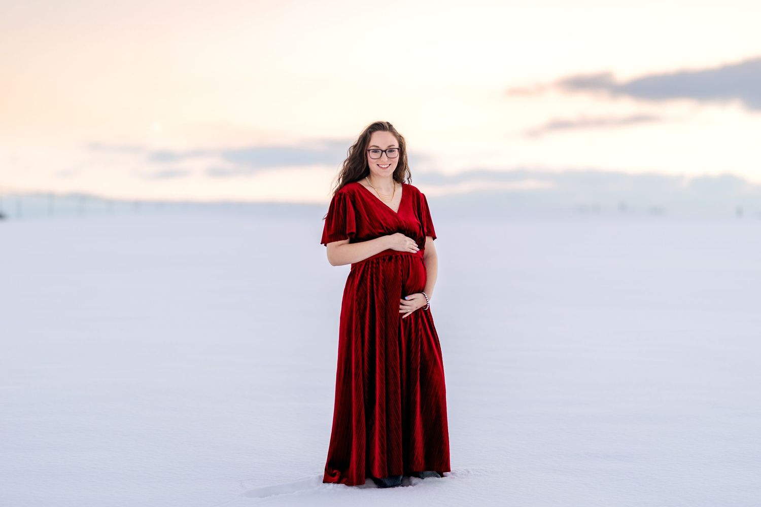 Smiling pregnant woman in a red velvet dress cradling her bump while standing in a snowy open field at sunset.