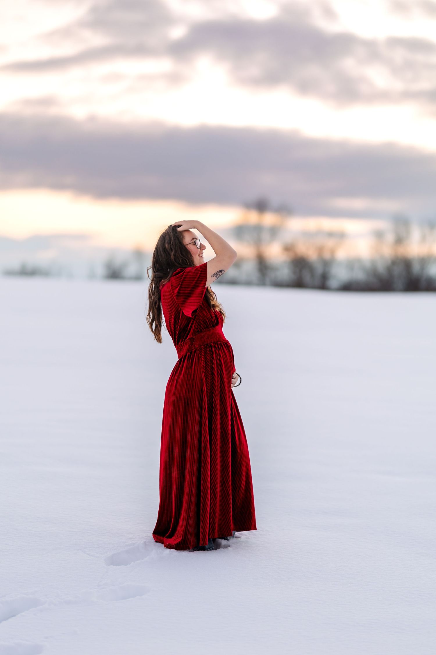 Pregnant woman in a flowing red velvet gown posing gracefully in a snowy field during a golden winter sunset.
