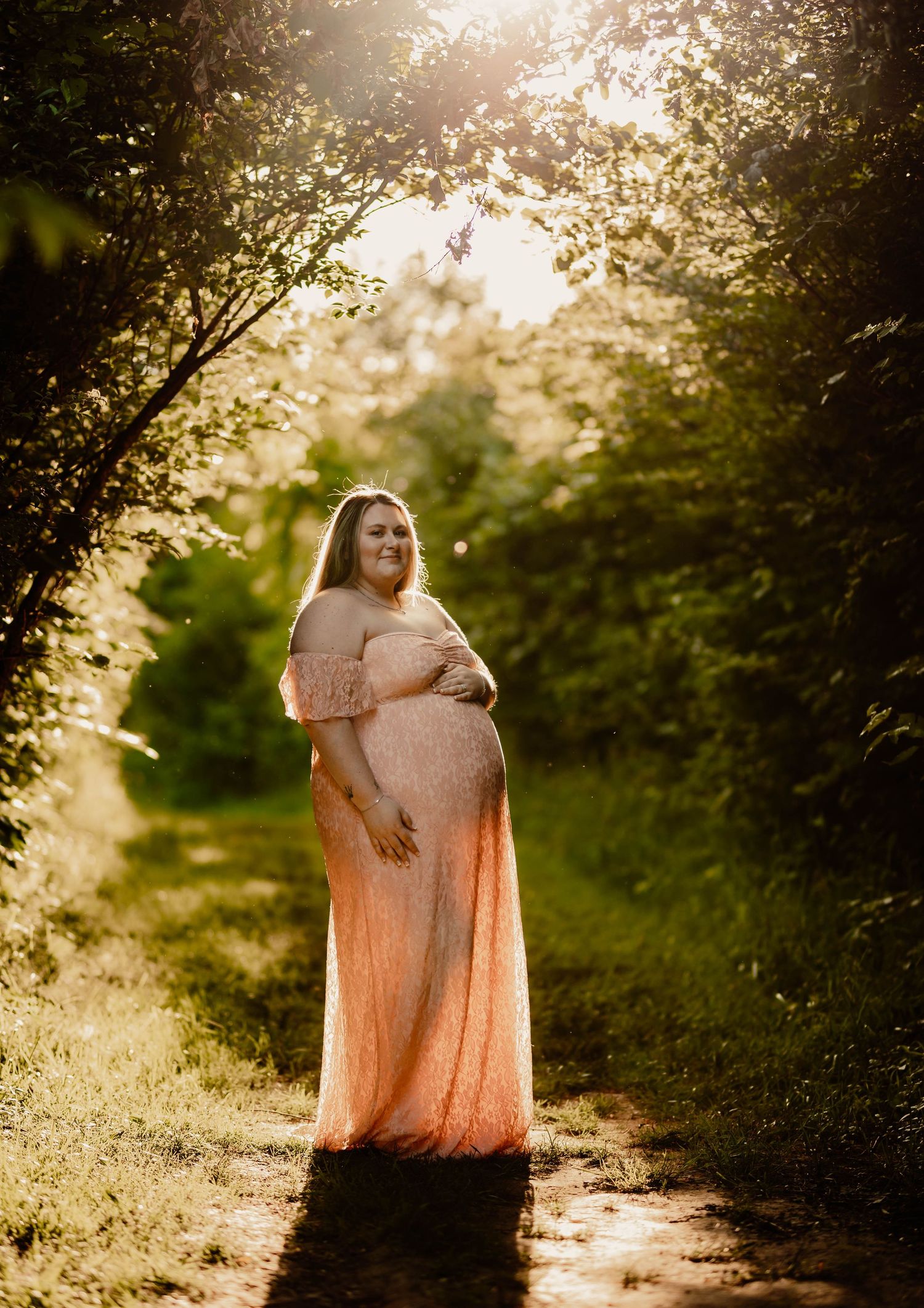 Pregnant woman in pink off-shoulder lace gown standing on sunlit forest path surrounded by green trees.