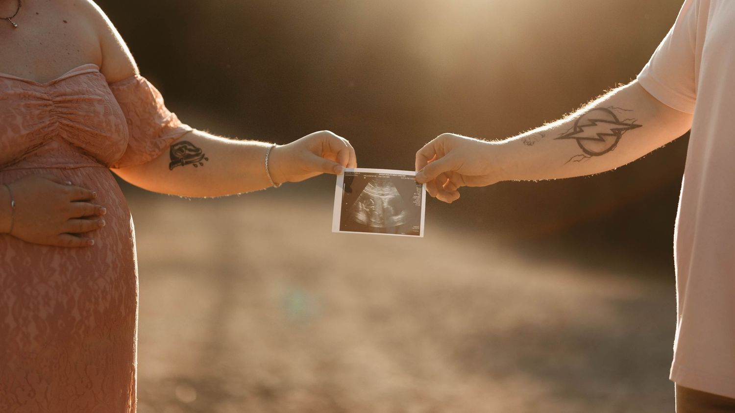 Expecting couple holding ultrasound photo together in golden sunlight, both showing tattoos on their arms.