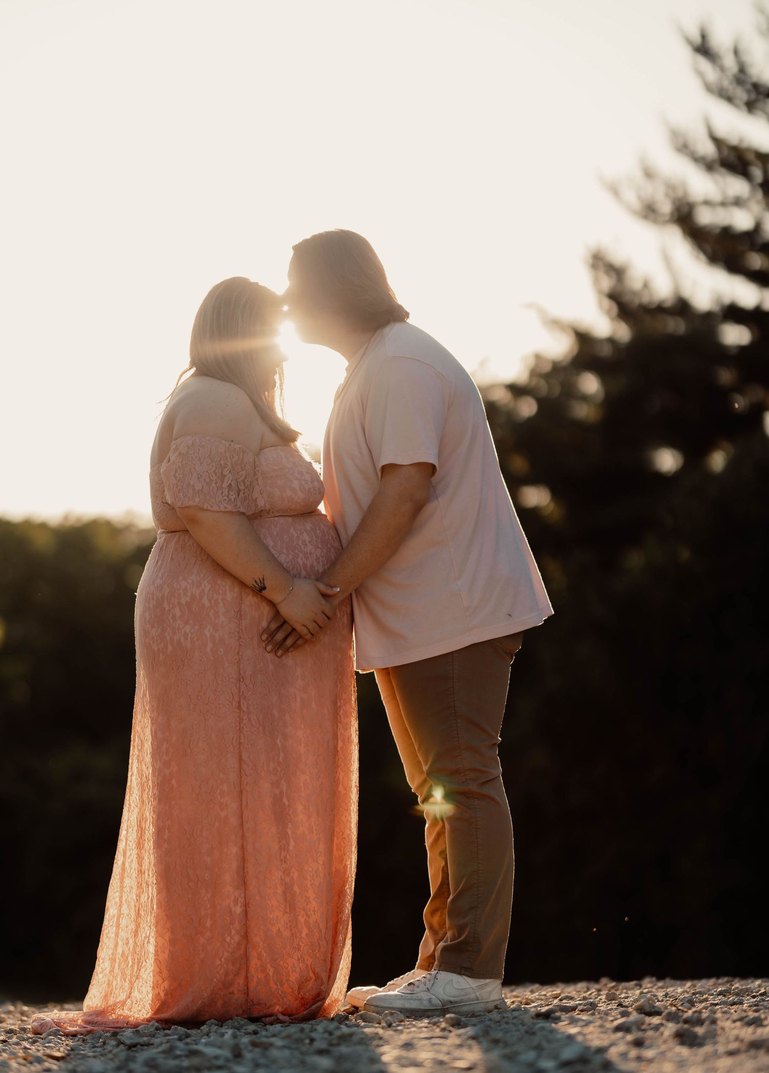 Pregnant woman in pink lace gown kissing partner at sunset with golden backlight during maternity photoshoot.