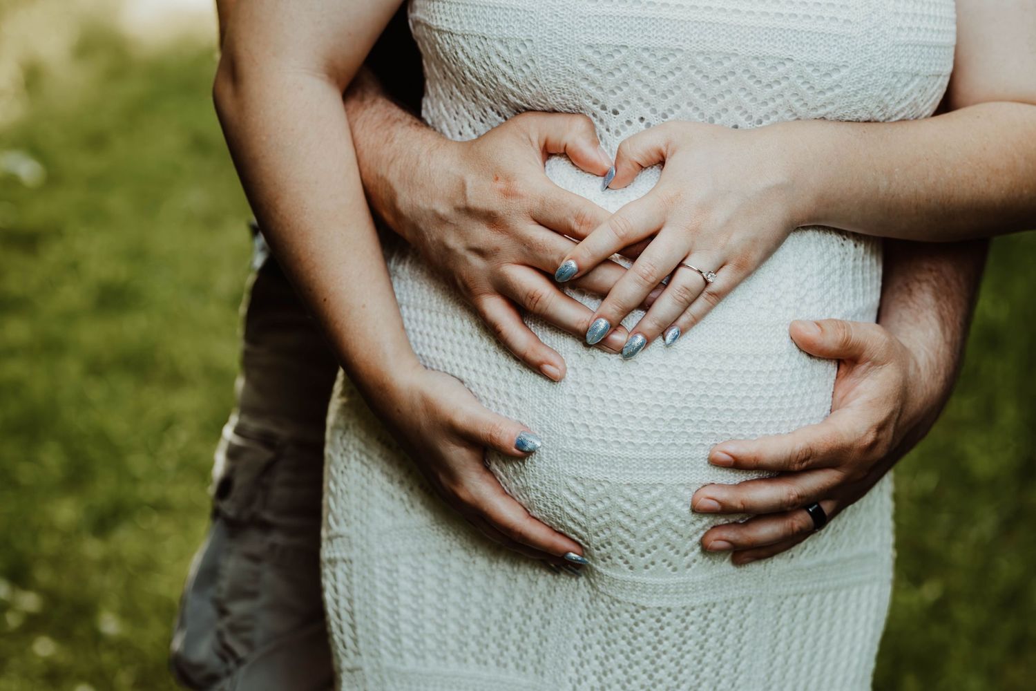Close-up of couple forming heart shape with hands over pregnant belly in white knit maternity dress.