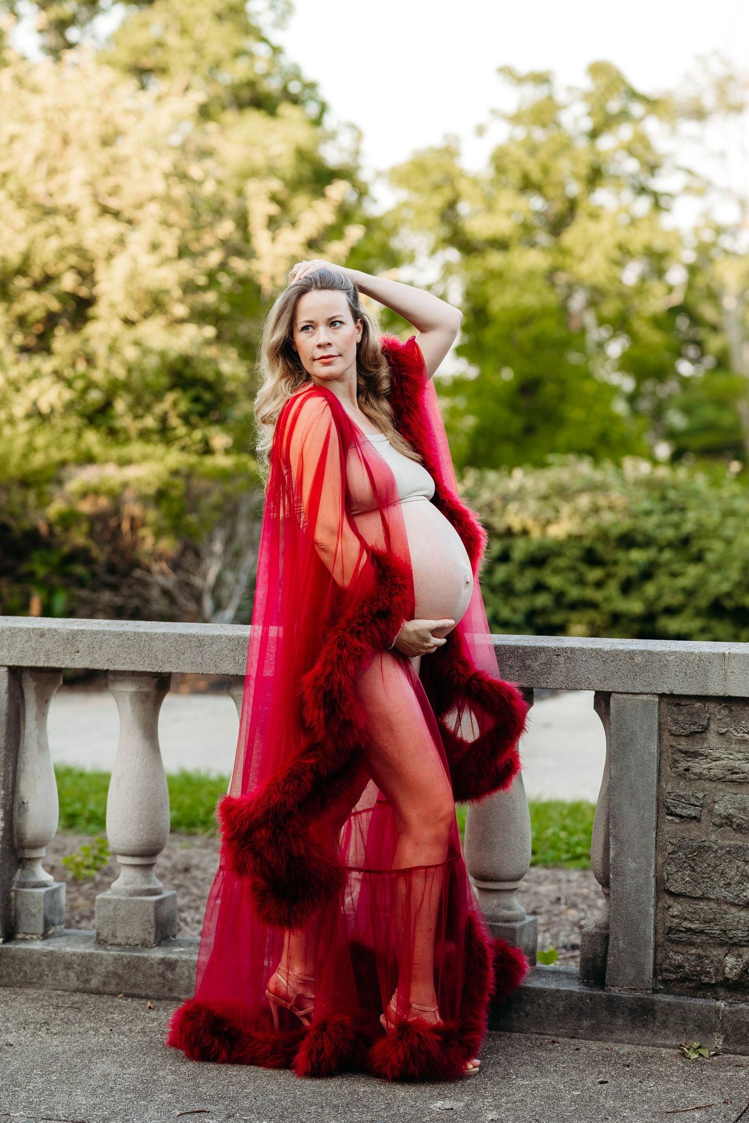Pregnant woman in sheer red feather-trimmed robe poses by stone balustrade outdoors.