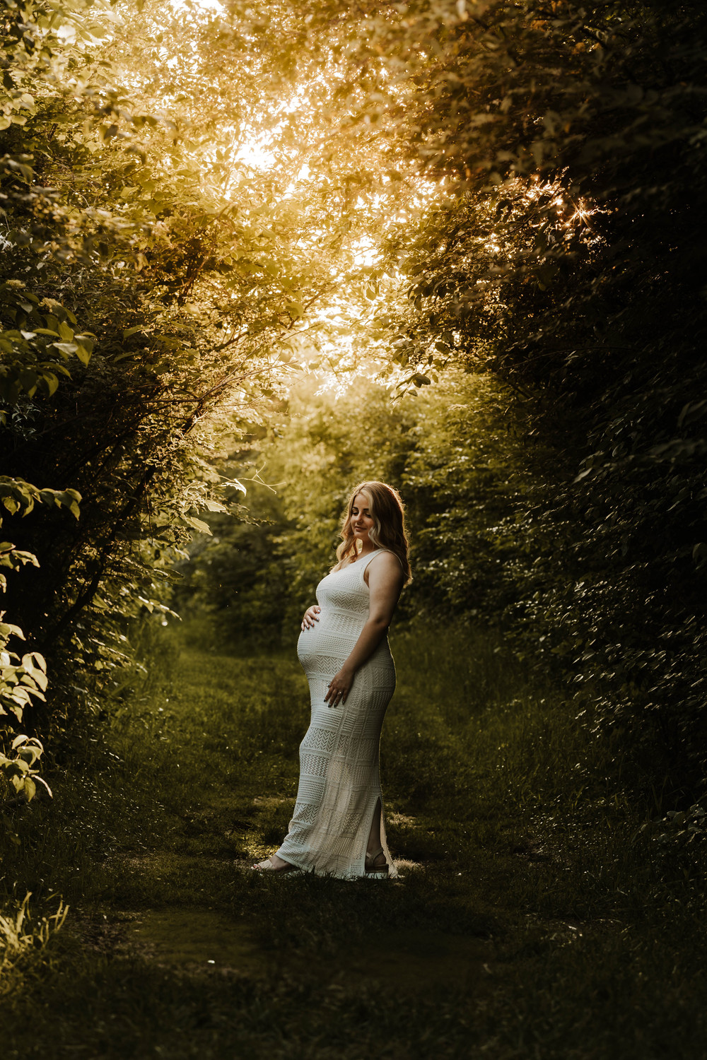 Pregnant woman in a white lace gown standing on a sunlit forest path surrounded by lush green trees.