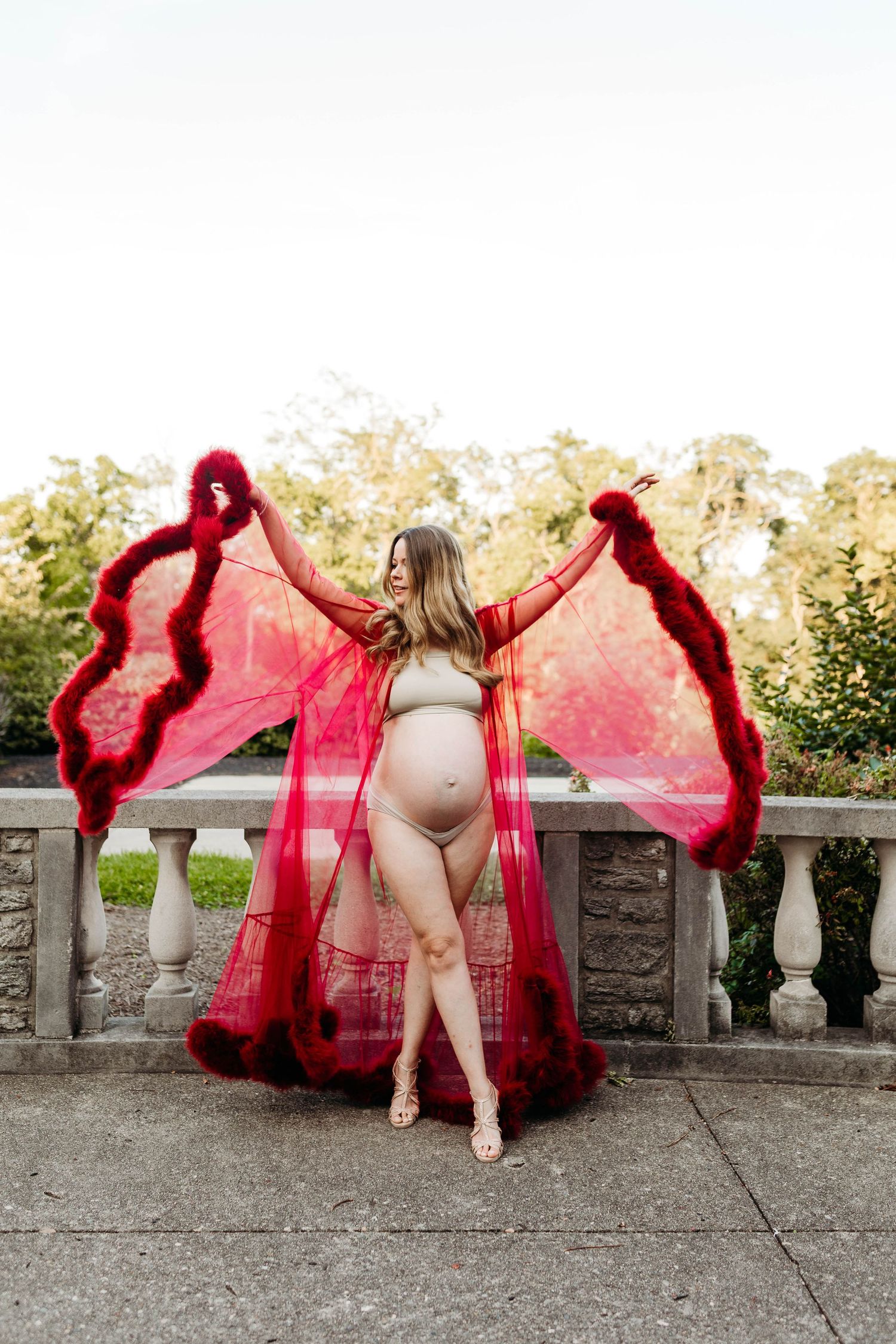 Pregnant woman spreads red feather-trimmed sheer robe like wings on outdoor stone balcony.