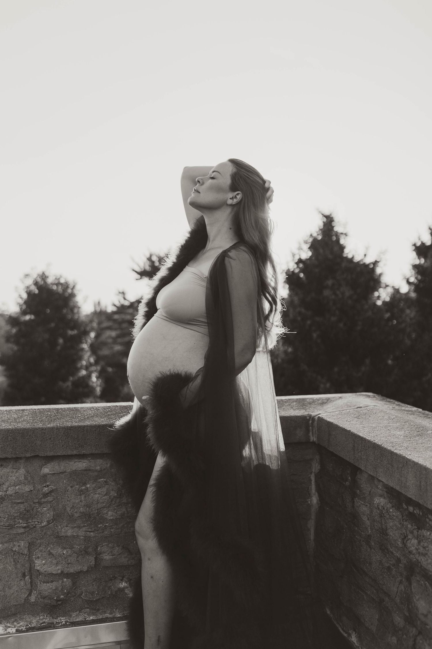 Black and white photo of pregnant woman in sheer robe gazing upward on stone balcony with trees.