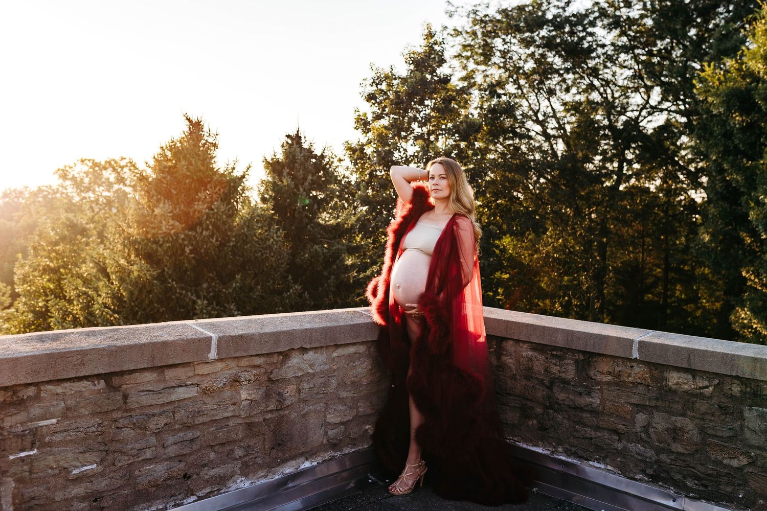 Pregnant woman in deep red flowing gown stands on stone terrace surrounded by golden sunset trees.