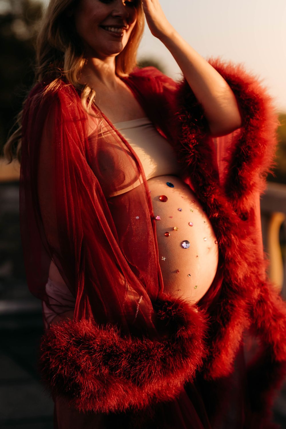 Close-up of pregnant belly adorned with colorful gems, woman wearing red feather-trimmed sheer robe.