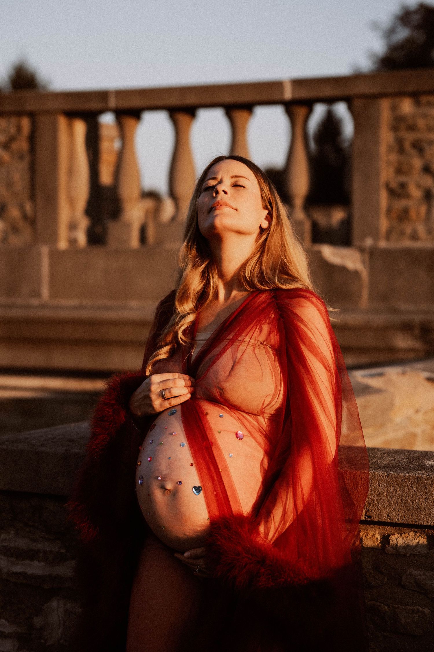 Pregnant woman in red sheer robe with gem-decorated belly basks in warm golden sunset light.