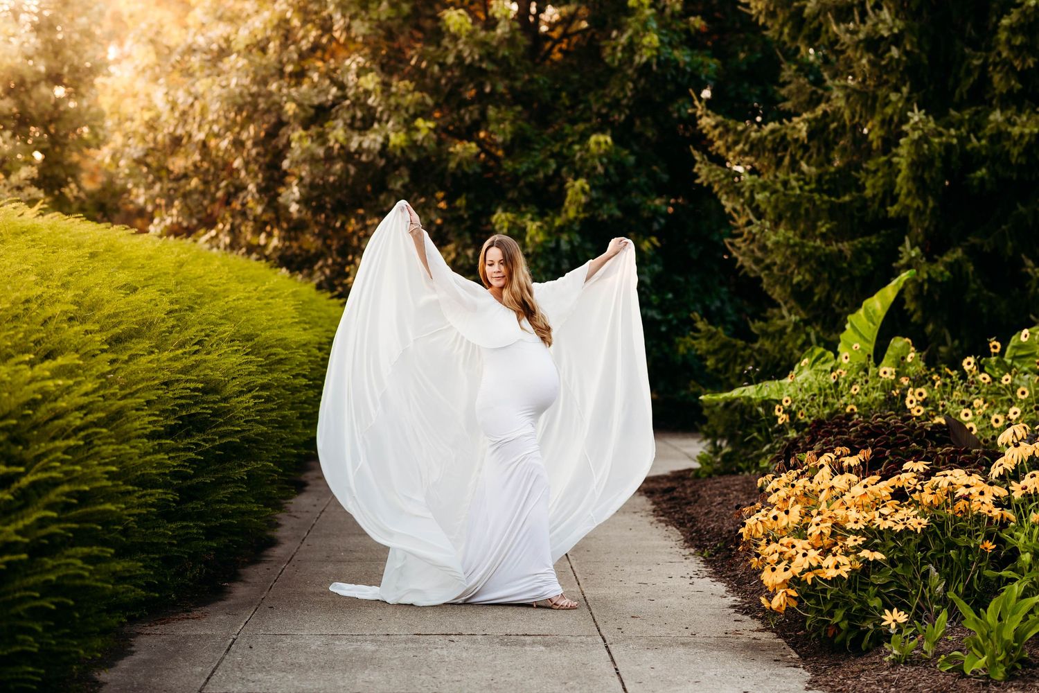 Pregnant woman in flowing white gown twirls on garden path surrounded by lush greenery and flowers.