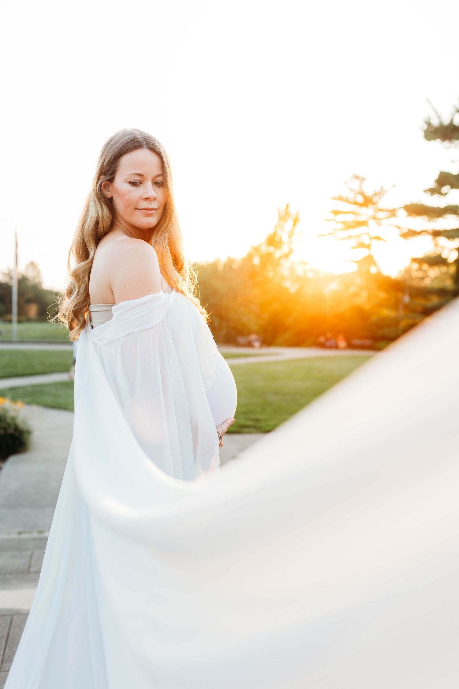 Pregnant woman in off-shoulder white dress holds flowing fabric in warm golden sunset light outdoors.