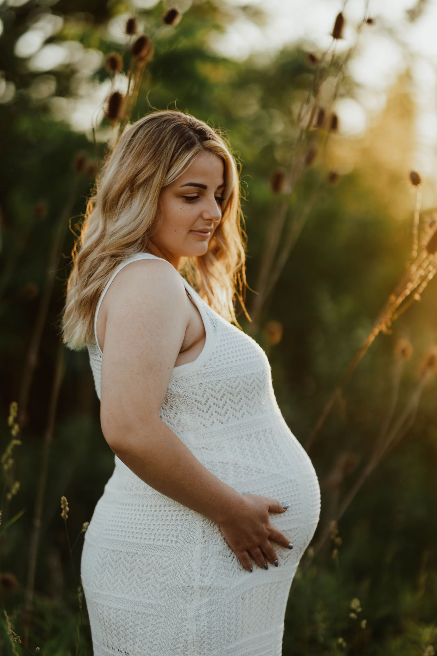 Blonde pregnant woman in a white lace dress cradling her bump in a golden sunlit meadow at sunset.