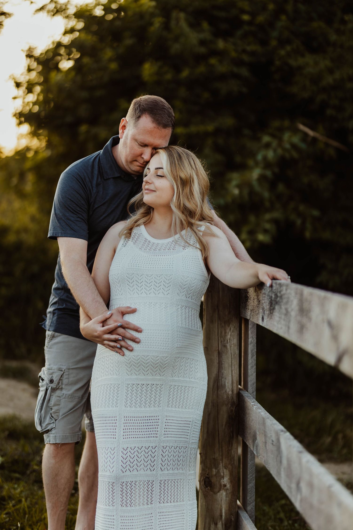 Pregnant woman in white knit dress with partner by wooden fence during golden hour maternity shoot.