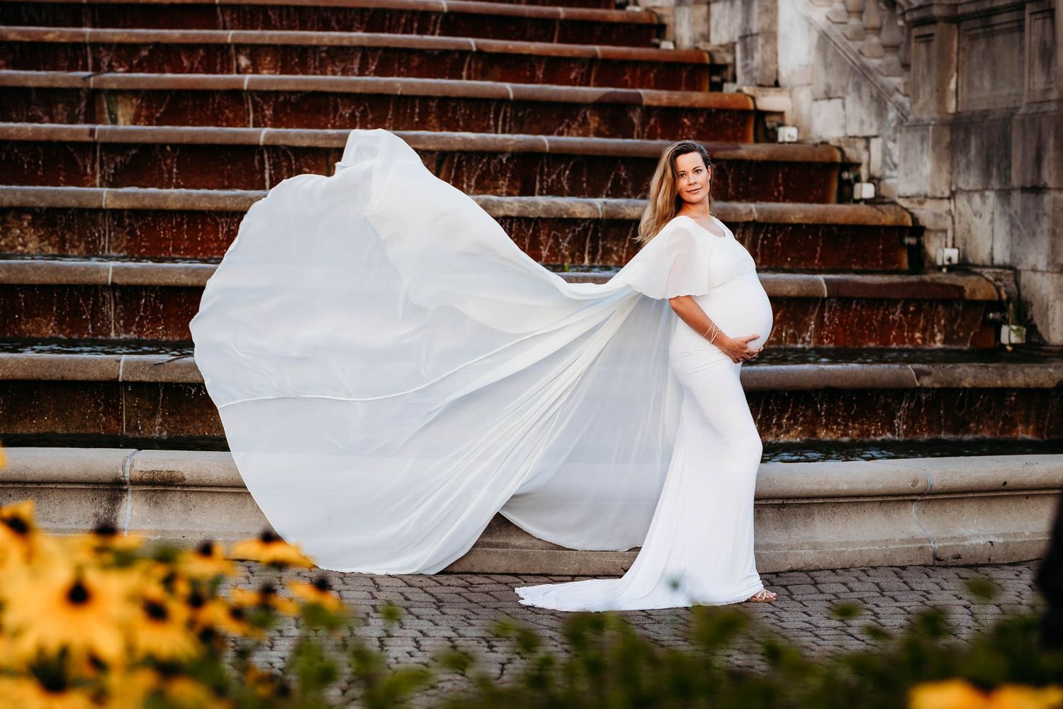 Pregnant woman in white flowing gown poses on stone steps with billowing fabric and yellow flowers.