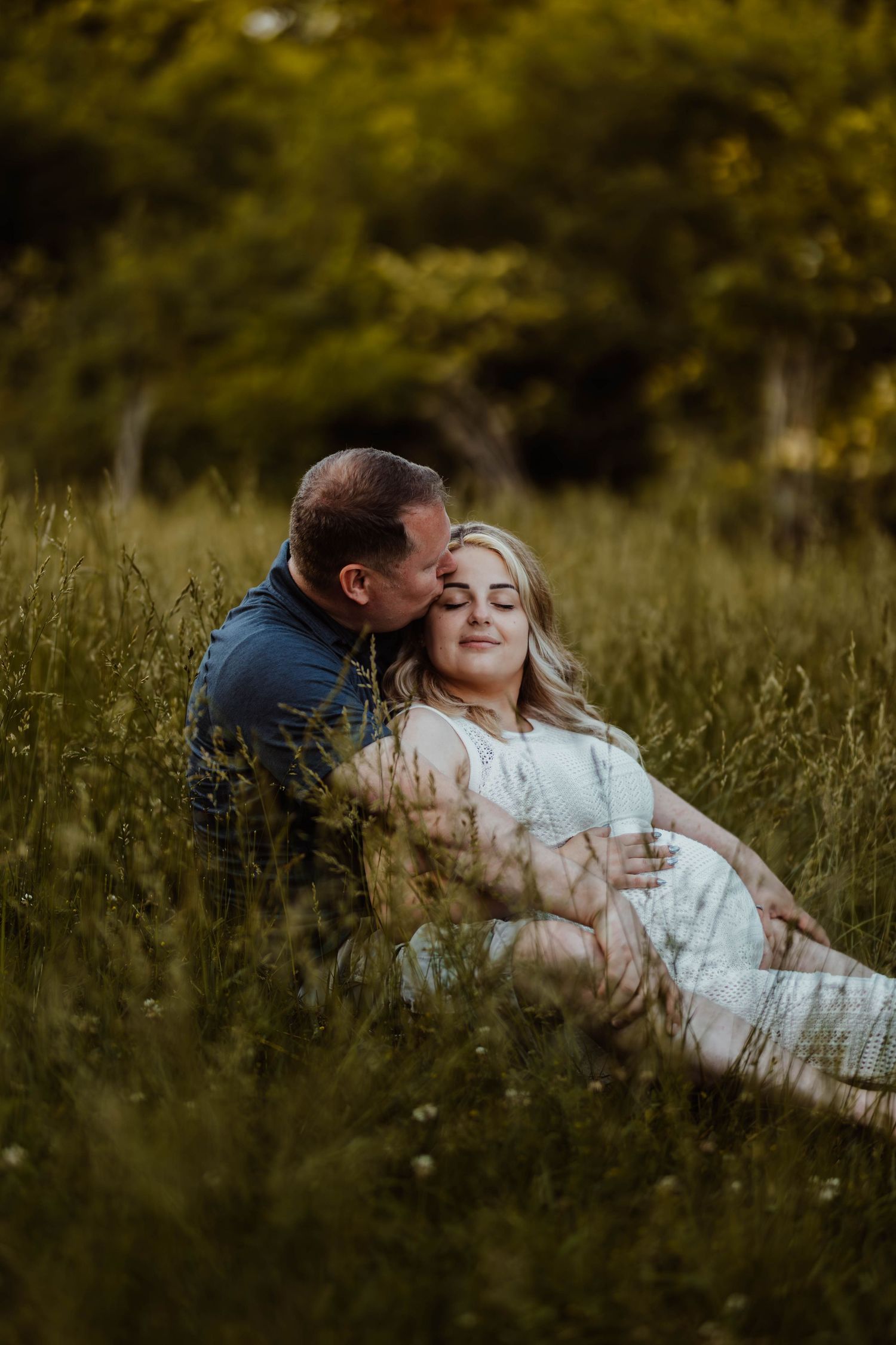 Couple sitting in tall green grass during a romantic outdoor maternity photography session at sunset.