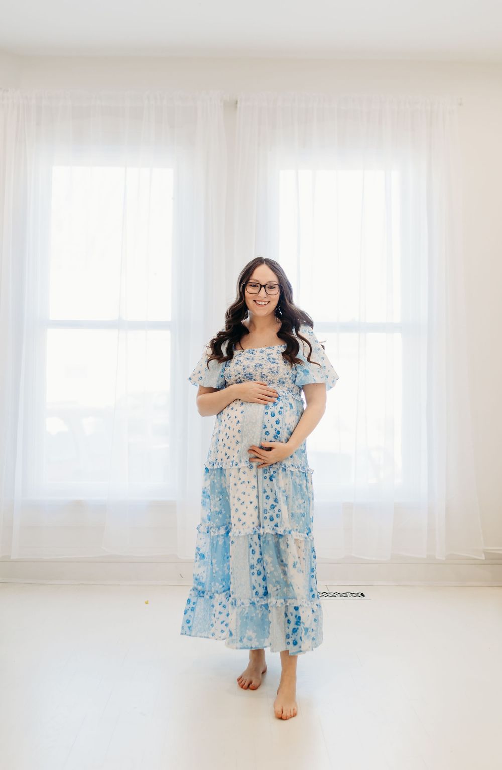 Smiling pregnant woman in a blue floral maternity dress cradling her baby bump in a bright white studio.