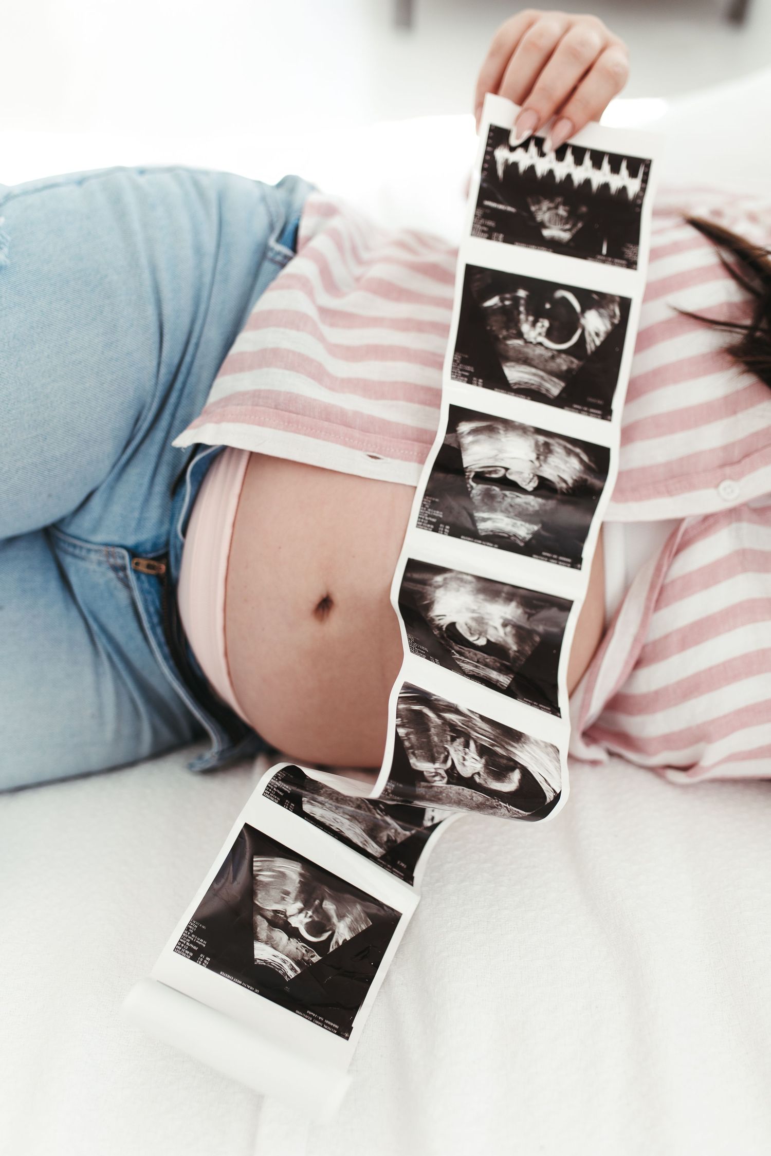 Pregnant woman lying down holding ultrasound strip over baby bump in striped shirt and jeans.