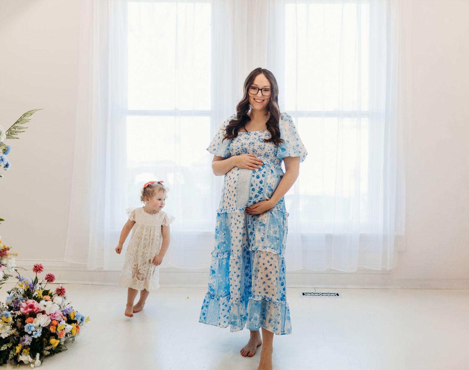 Pregnant woman in blue floral dress holding baby bump while toddler walks nearby among flowers in white studio.