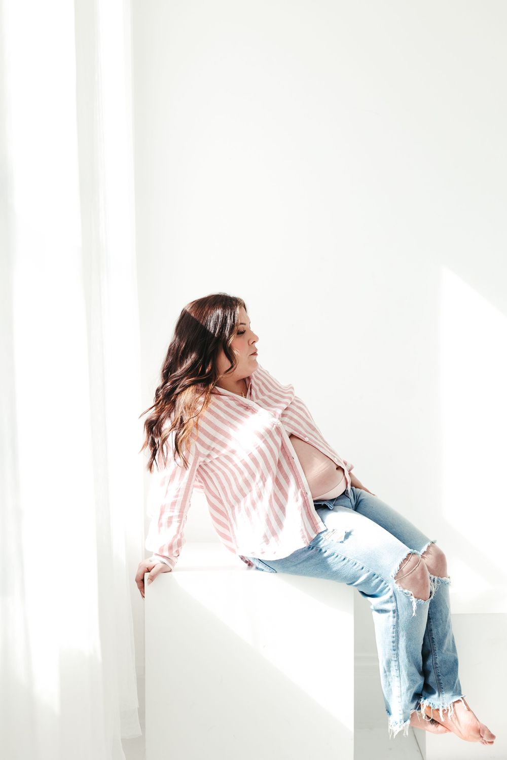 Pregnant woman in pink striped shirt and ripped jeans sitting in bright sunlit white studio.