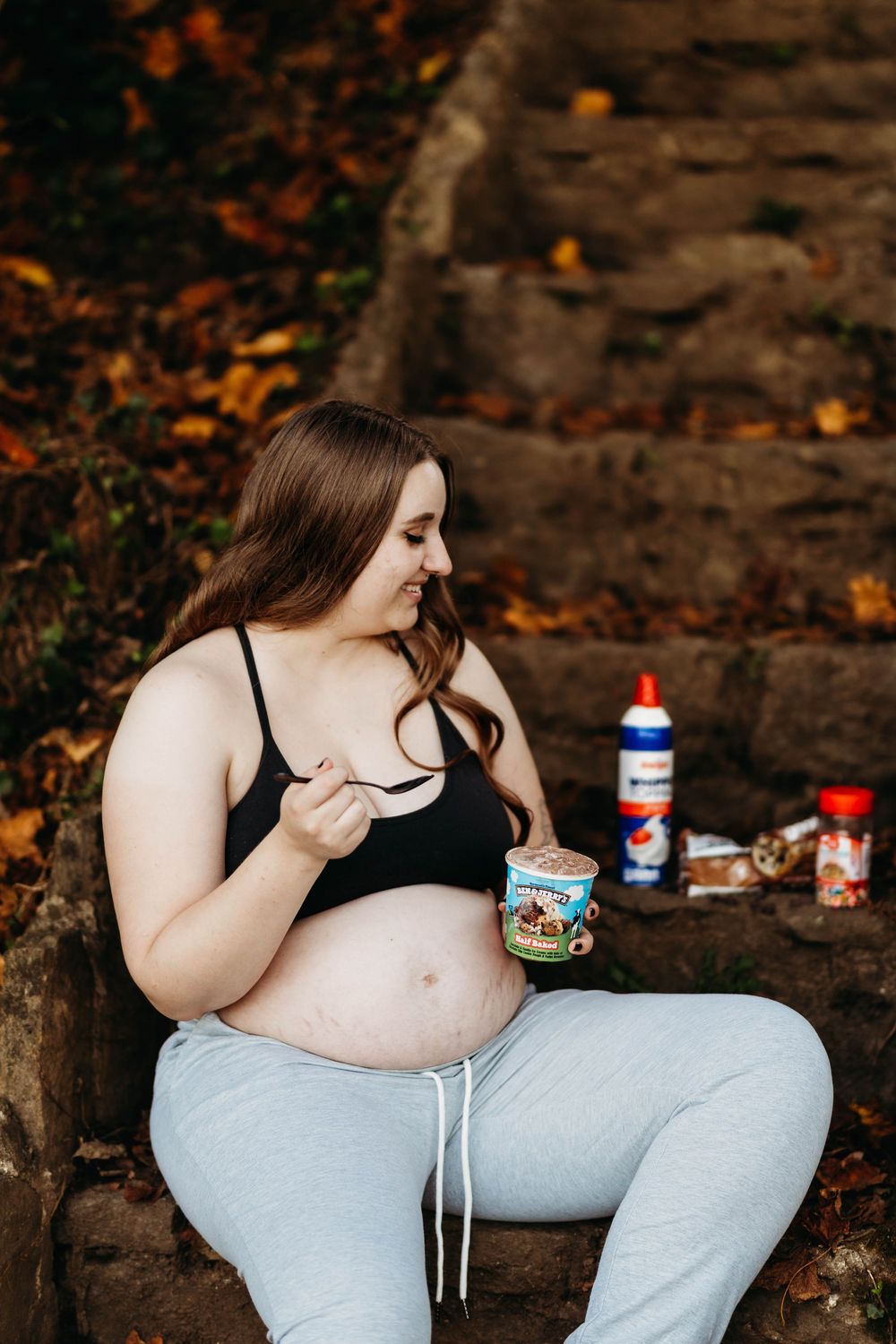 Smiling pregnant woman sits on stone steps eating ice cream surrounded by autumn leaves outdoors.