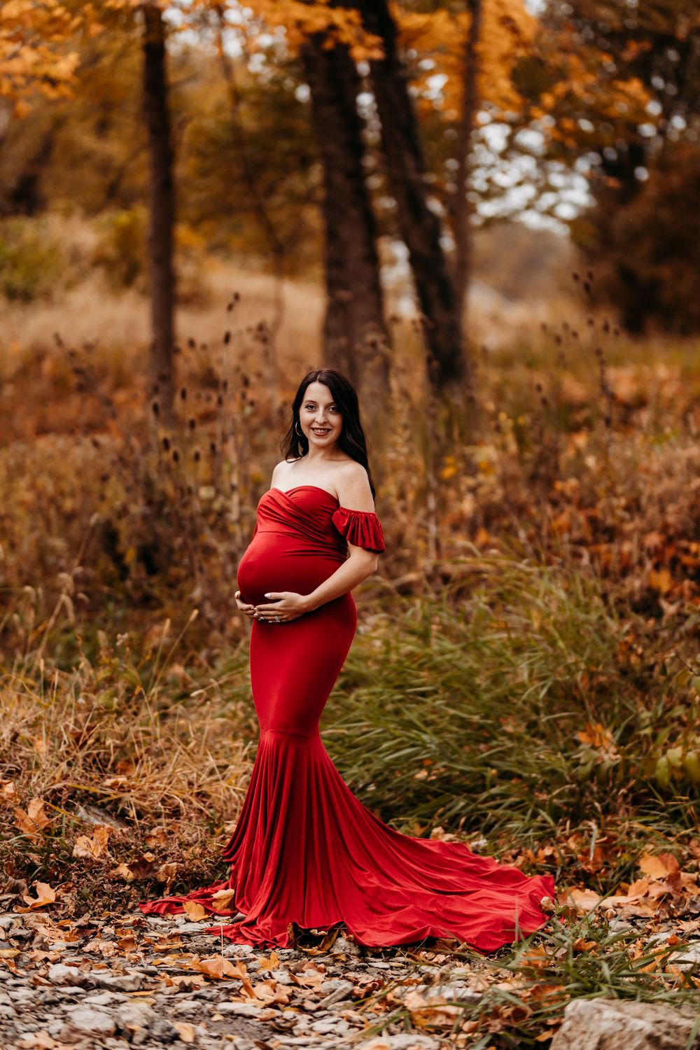 Smiling pregnant woman in red off-shoulder mermaid gown stands among autumn trees and fallen leaves outdoors.