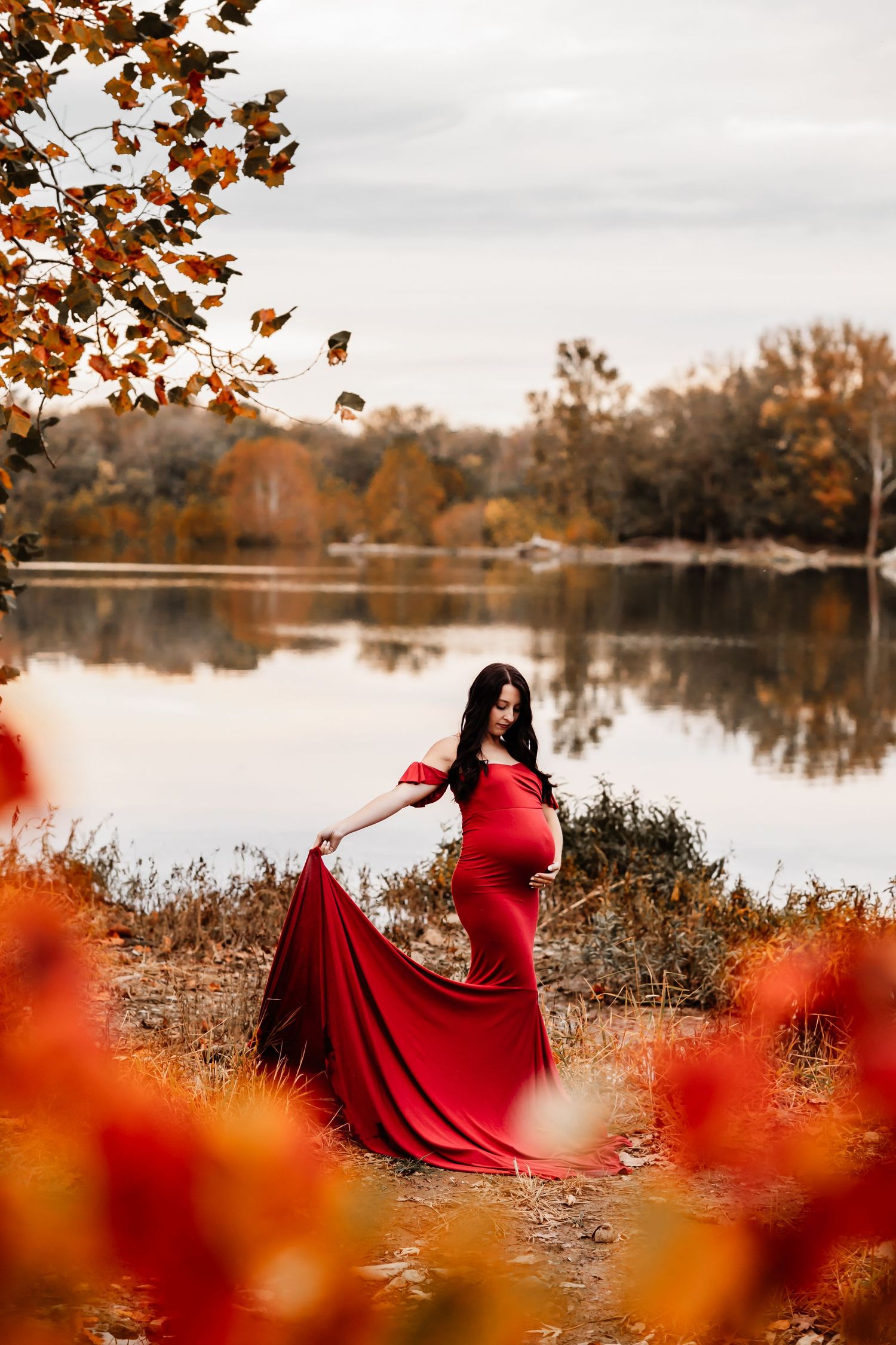 Pregnant woman in red off-shoulder gown with train poses by autumn lake surrounded by colorful fall foliage.