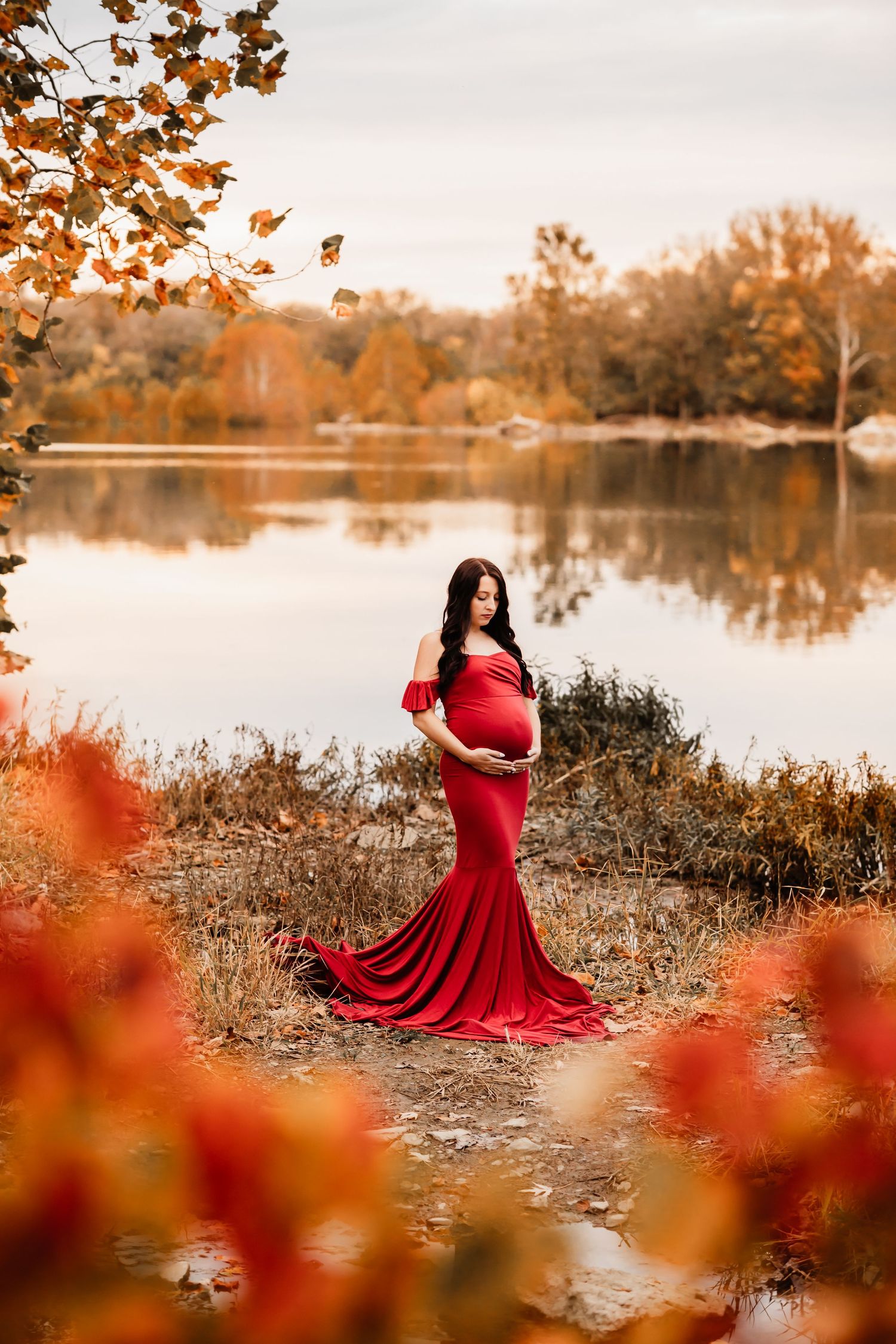 Pregnant woman in elegant red gown with train cradles bump beside serene autumn lake with colorful foliage.
