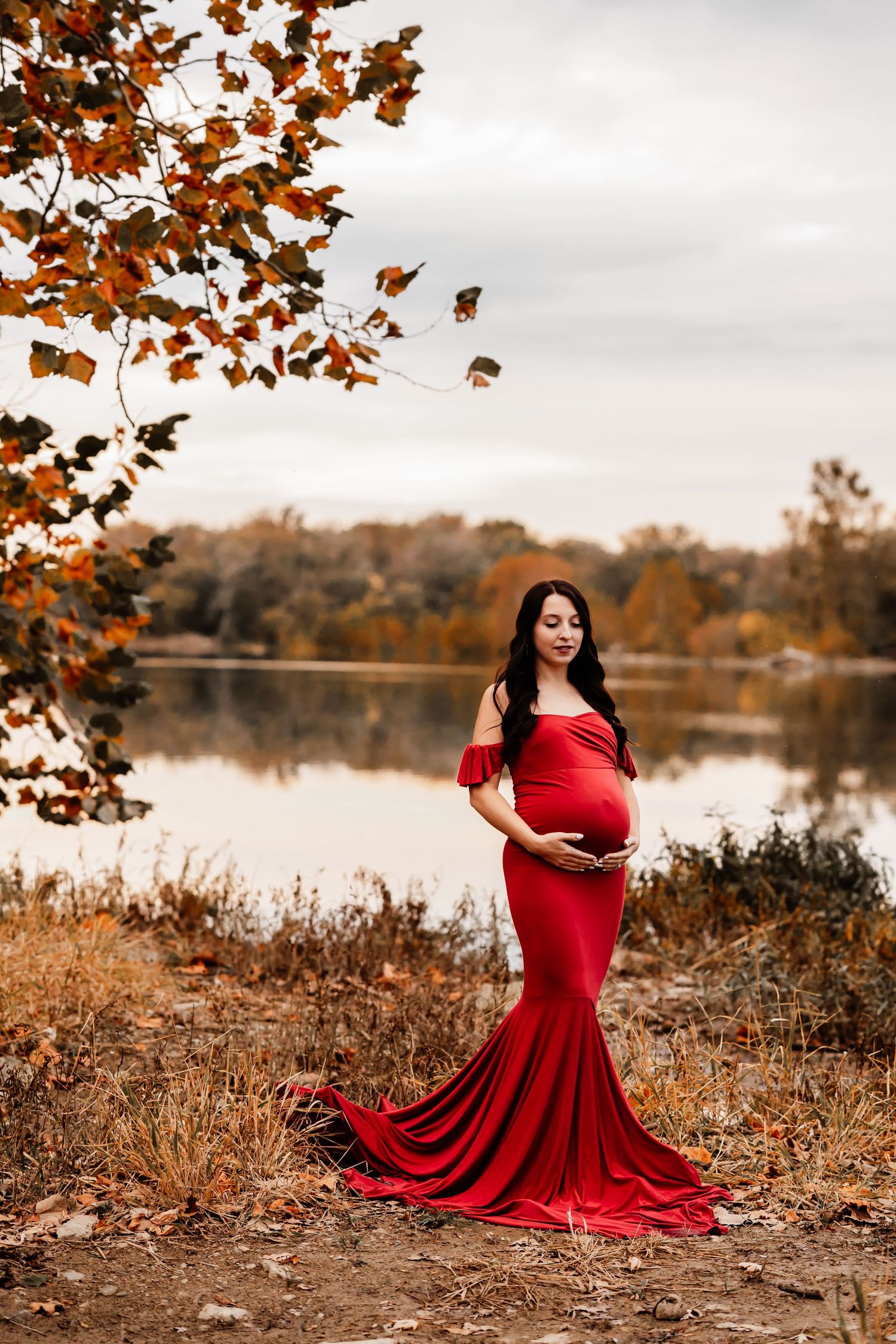 Pregnant woman in red off-shoulder gown with long train poses by autumn lake under colorful fall leaves.