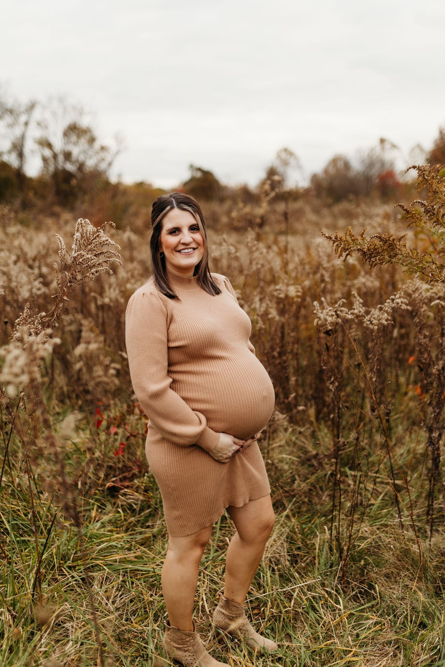 A pregnant woman in a tan ribbed dress smiles while holding her belly in a dry autumn meadow.