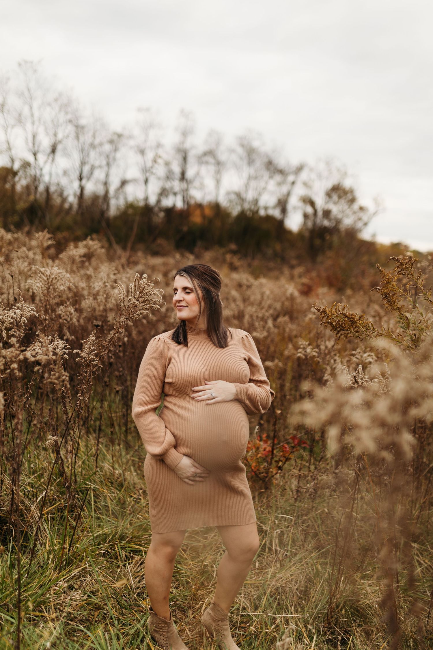 A pregnant woman in a camel-colored dress gently holds her bump while standing among tall autumn grasses.