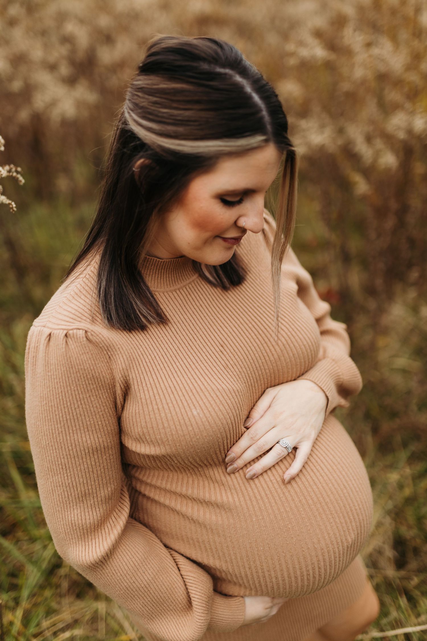 A pregnant woman in a tan ribbed sweater dress lovingly cradles her belly while looking down outdoors.