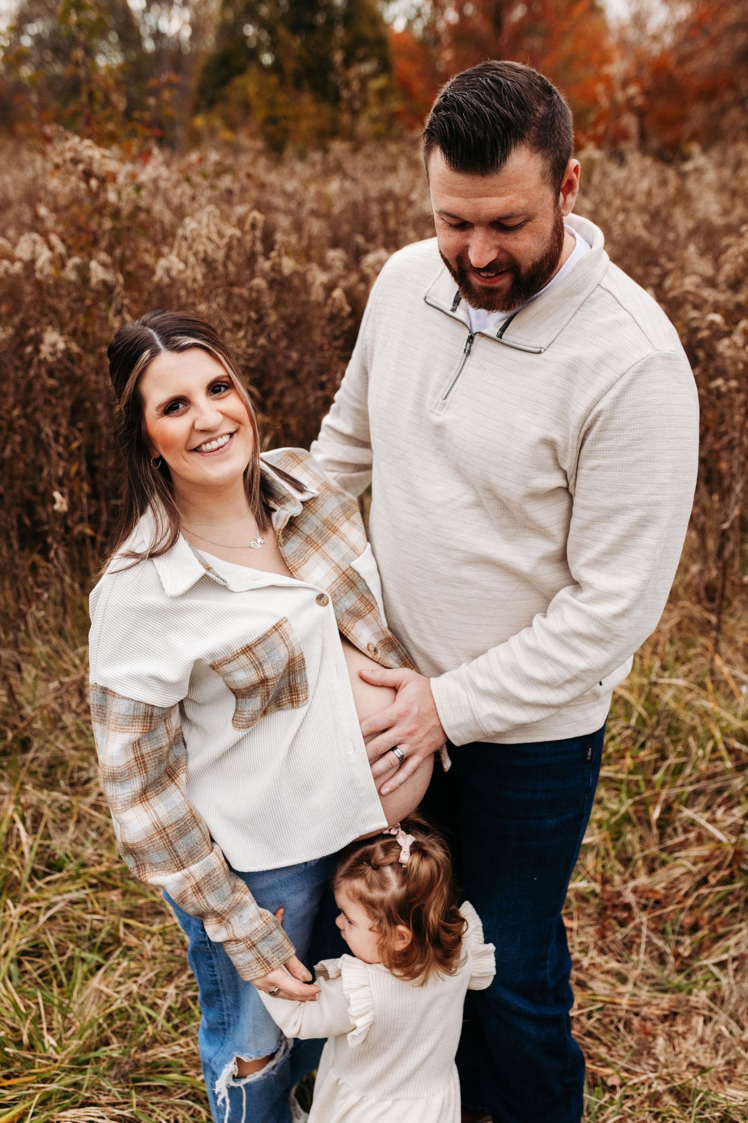 A pregnant couple smiles as their young daughter touches mom's baby bump in a fall outdoor setting.