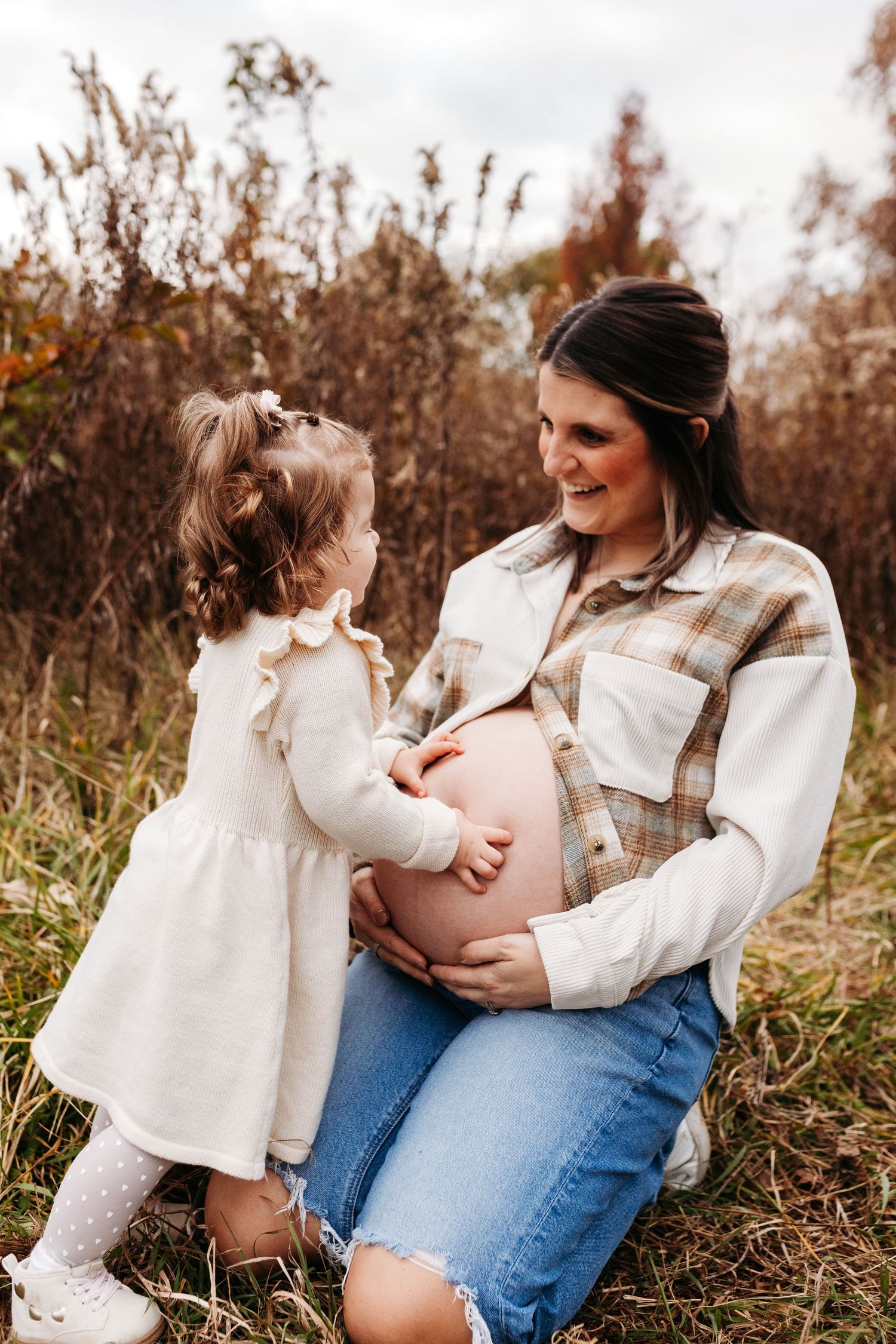 A pregnant woman sits outdoors as her young daughter gently touches her bare baby bump in autumn.