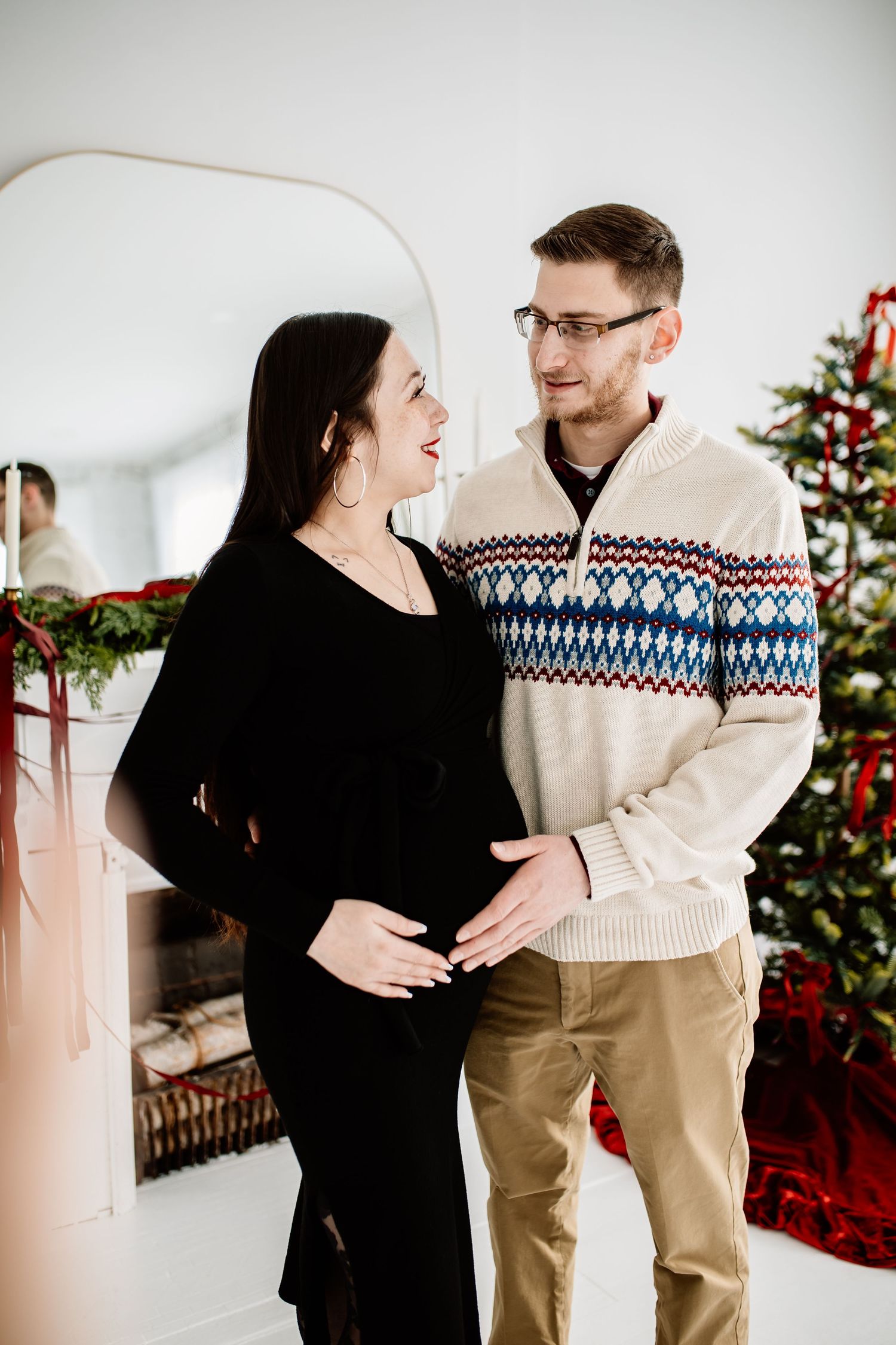 Smiling couple in holiday sweaters cradle pregnant belly near decorated Christmas tree indoors.