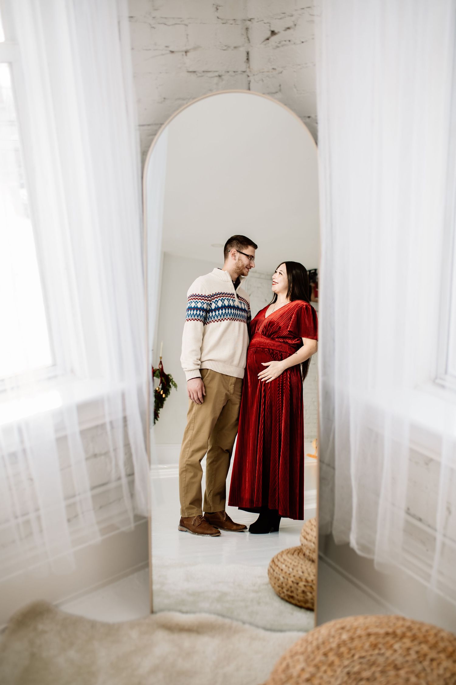 Couple poses in mirror reflection, pregnant woman in red velvet dress with partner in fair isle sweater in white room.