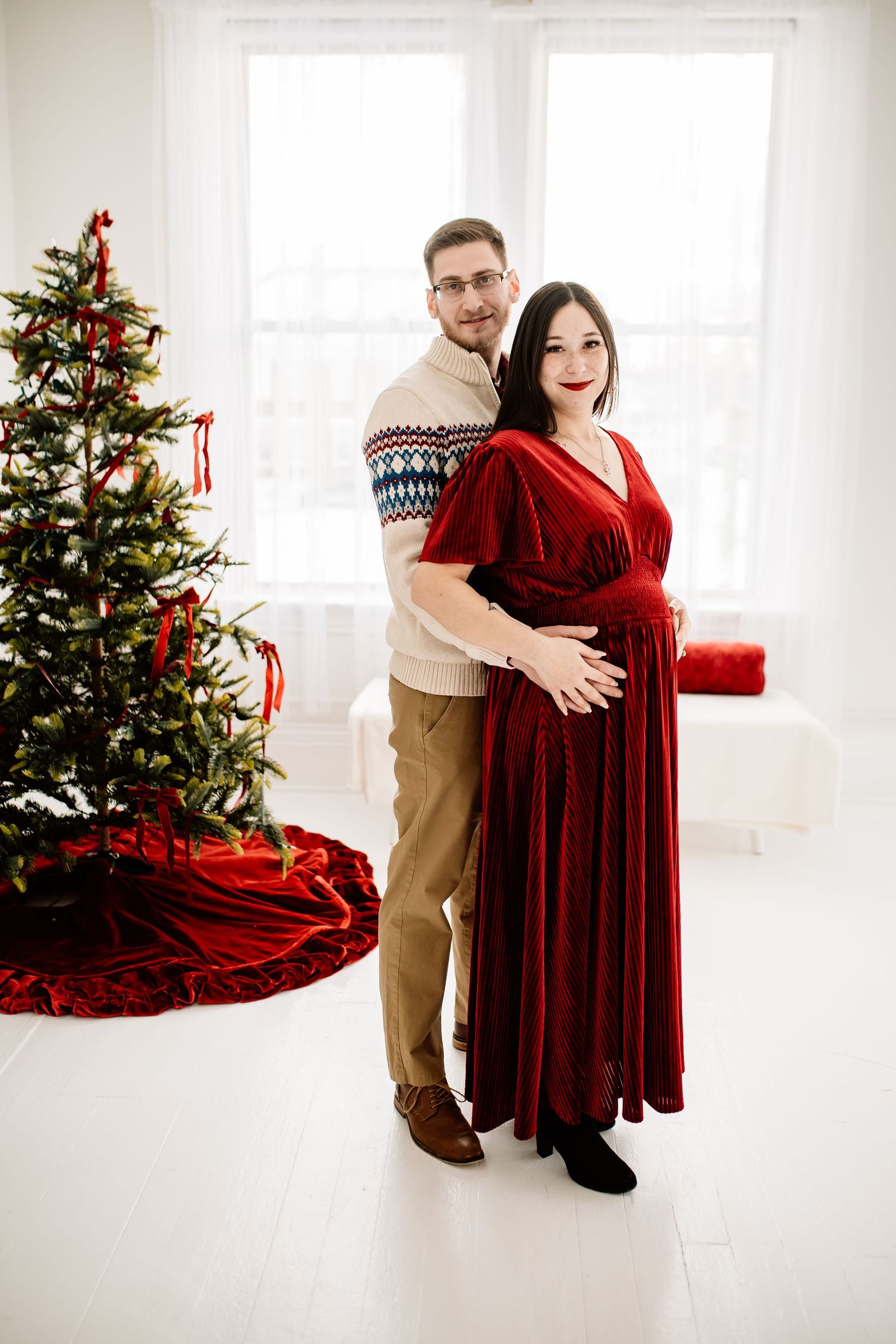 Pregnant woman in red velvet dress with partner embracing her beside decorated Christmas tree in bright white room.