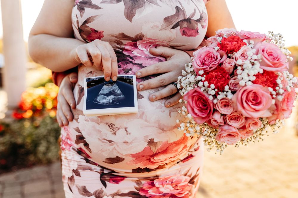 Pregnant woman in floral dress holding ultrasound photo and pink rose bouquet during maternity shoot.