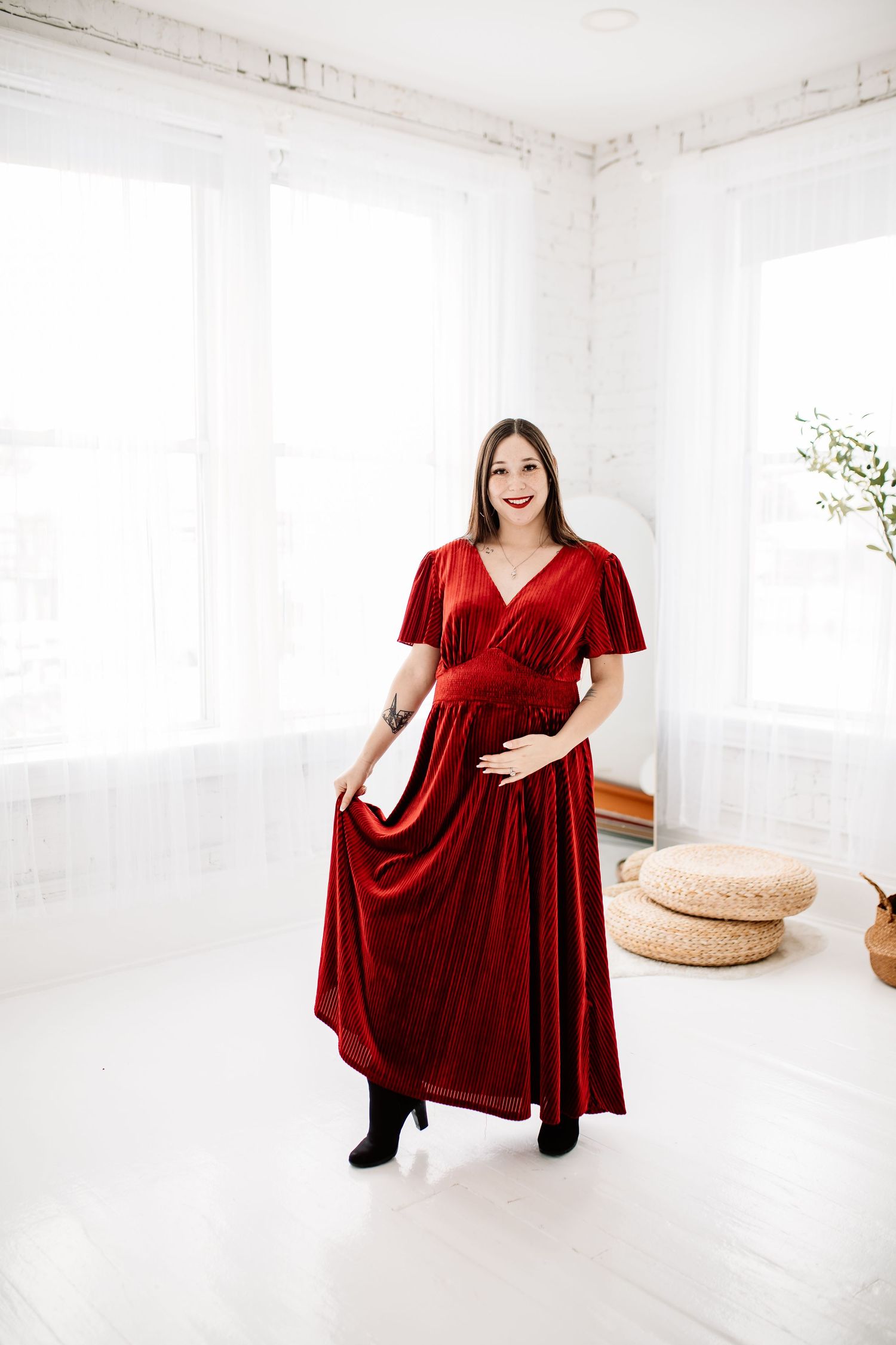 Smiling pregnant woman in flowing red velvet dress poses in bright white studio holding her baby bump.