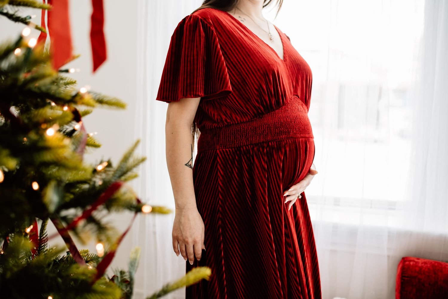 Close-up of pregnant woman in red velvet dress cradling baby bump beside decorated Christmas tree.