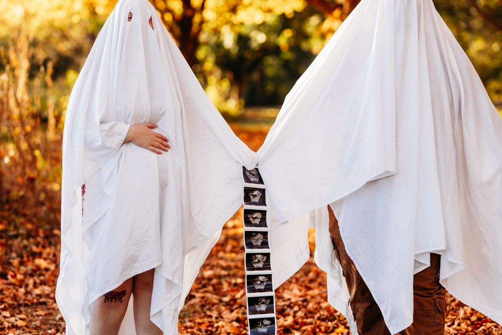 Pregnant couple dressed as ghosts in white sheets holding ultrasound photos in autumn park.