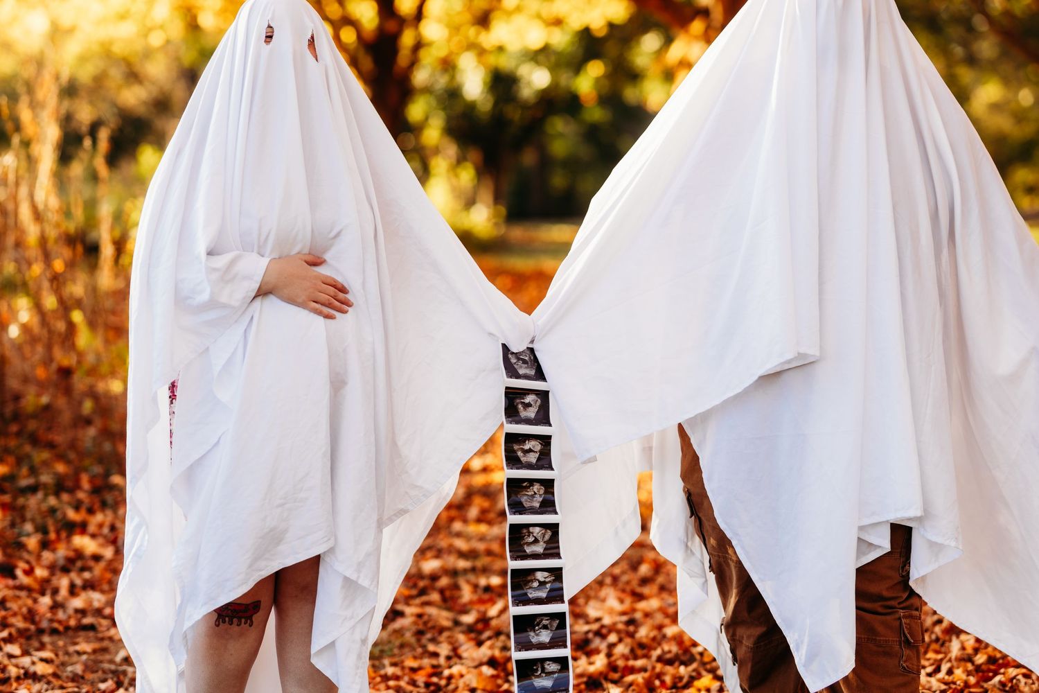 Pregnant couple dressed as ghosts in white sheets holding ultrasound photos in autumn park.