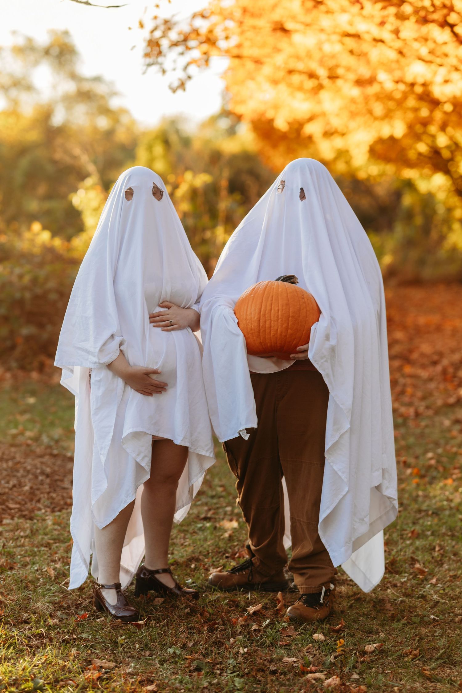 Couple in ghost costumes with pregnant belly and pumpkin in fall foliage outdoor setting.