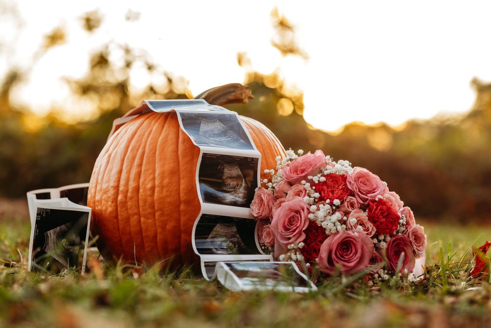 Baby ultrasound images and pink floral bouquet beside pumpkin in golden sunset light.