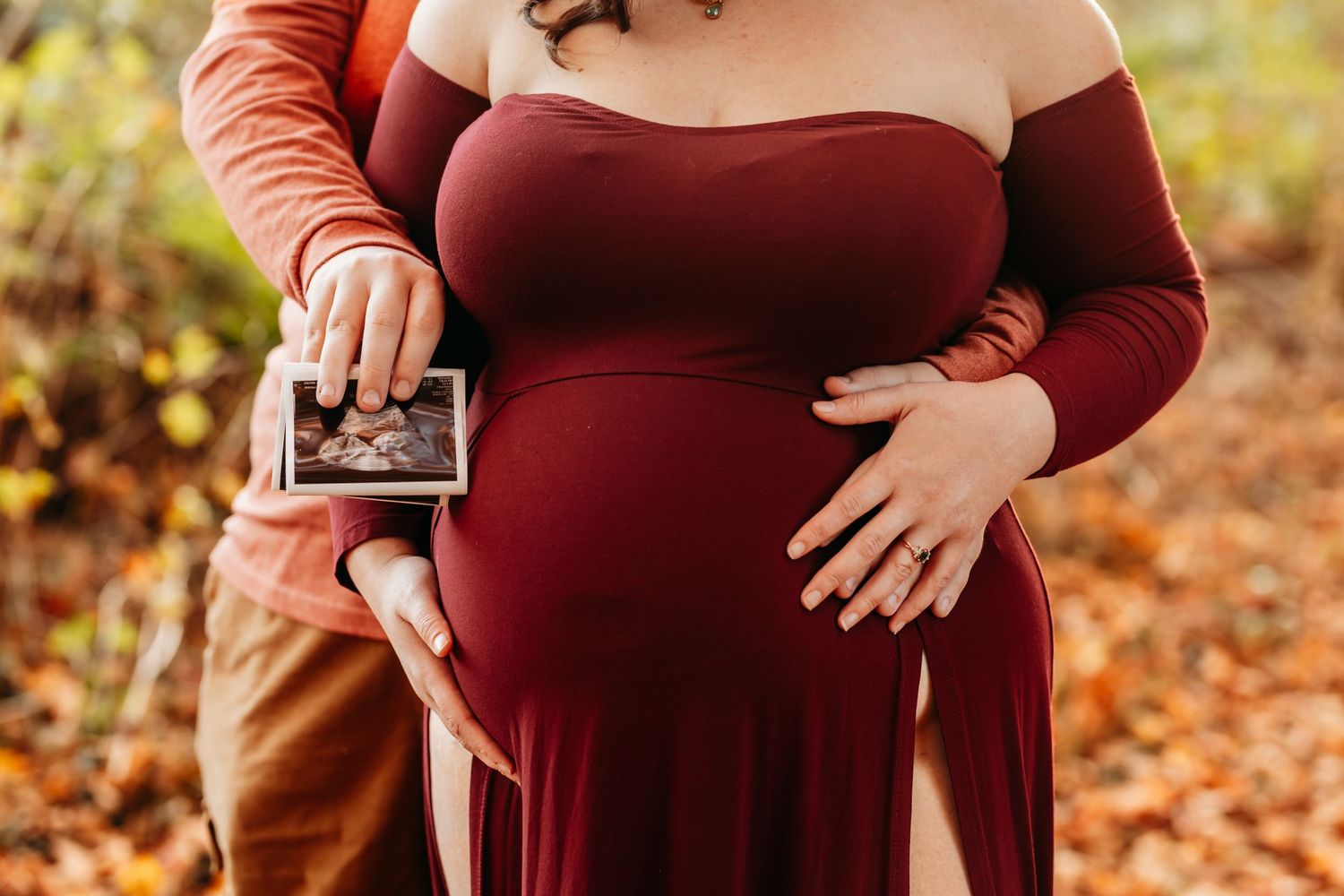 Pregnant woman in burgundy dress holding ultrasound photo with partner in fall forest.