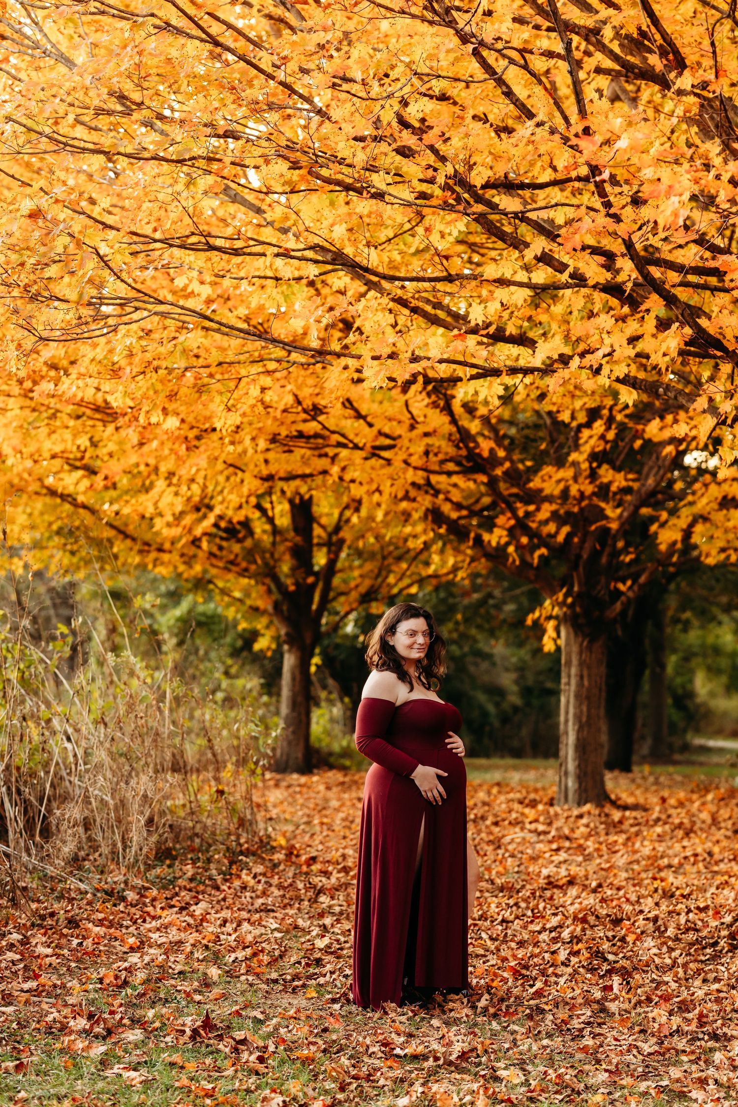 Pregnant woman in burgundy off-shoulder gown standing under golden autumn trees in park.