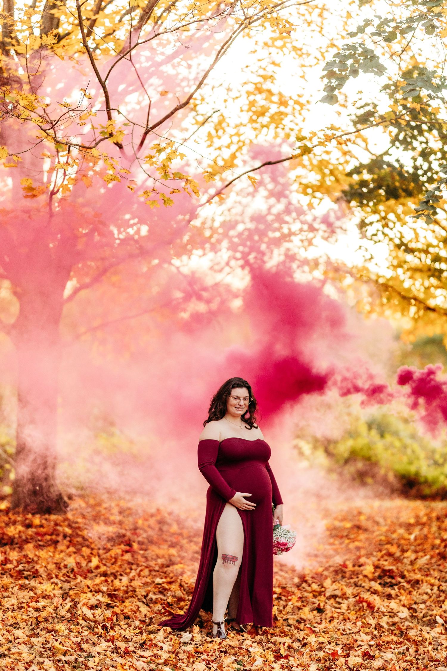 Smiling pregnant woman in red gown surrounded by pink smoke bomb in fall foliage.