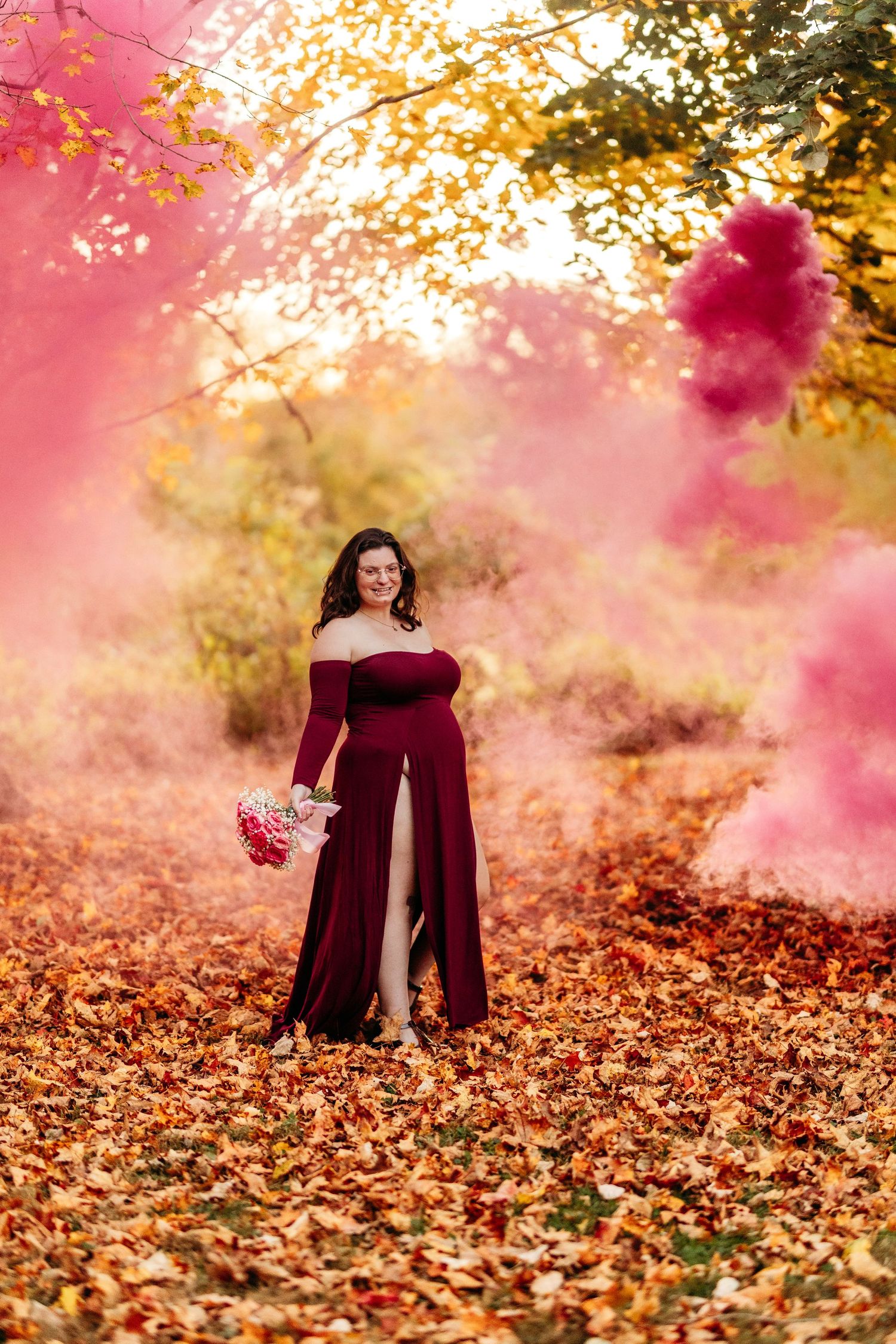 Pregnant woman in red gown holding bouquet amid pink smoke bombs in golden fall forest.