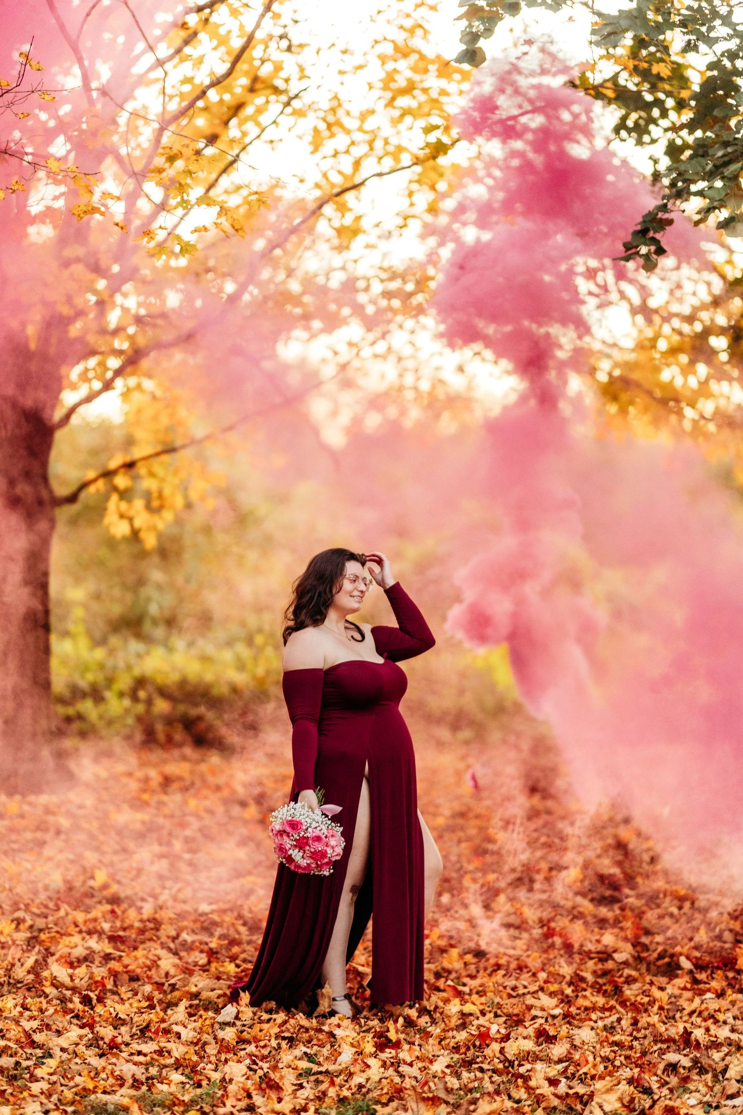Glowing pregnant woman in burgundy dress posing with bouquet in pink smoke autumn setting.