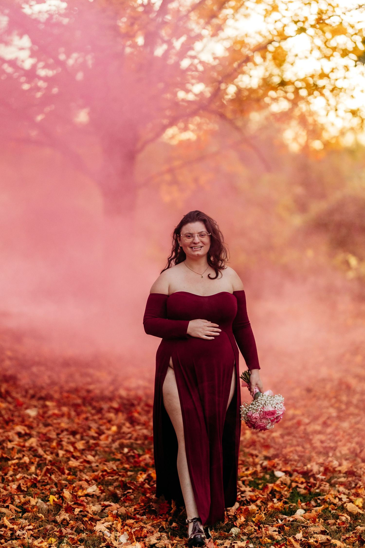 Pregnant woman in burgundy dress walking through pink smoke with bouquet in autumn leaves.